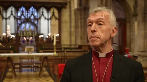 Andrew John in a red shirt and black blazer with a dog collar, looking at the camera in front of an altar. He has grey hair and brown eyes