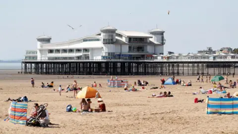 Getty Images Families sit on a beach on a sunny day with a long pier in the background