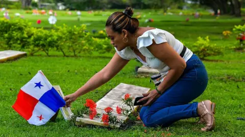 Getty Images A young woman in white top, blue jeans and sandals puts a Panamanian national flag on a grave in a neatly kept cemetery.