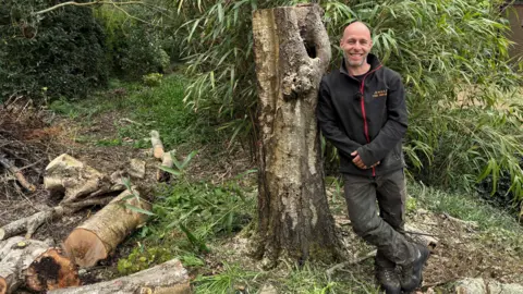 Pete Waterfield leaning against a tree stump after the tree has been chopped in half. Waterfield is wearing black combat style trousers and a black all-weather jacket smiling at the camera in a wooded area.