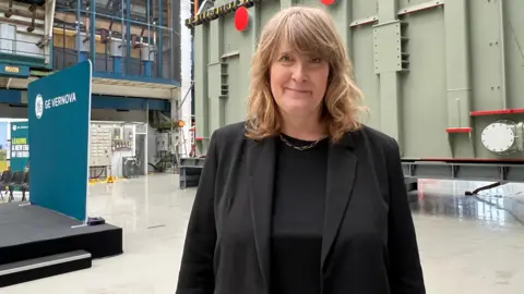 A woman with long blonde hair and a black top and jacket, stands and smiles in the middle of a large factory room.