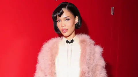 Nara Smith wears a pink fluffy cardigan on top of a silky white shirt which has a black bow on the front. She has a short cropped black haircut and wears light pink lipstick