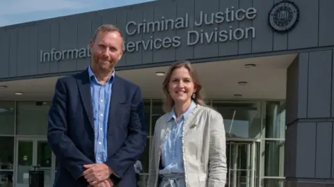 Zoe Billings A man and a woman stand in business wear in front of a grey, modern building. The front on the building reads 'Criminal Justice Information Services Division' next to the FBI logo. 