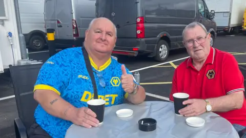A man on the left is wearing a mainly blue top and the man on the right is in a red top - they both include a Wolves logo. The men are sitting down at a table and both holding a drinks cup. 