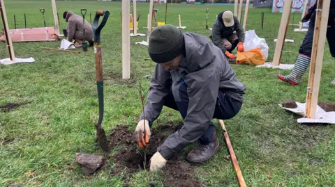 A man crouches down and plants a sapling in a hole he has dug. He is surrounded by other people also planting trees. 