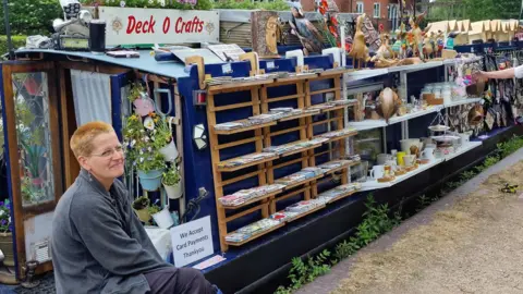 A woman sits at the end of a canal boat will shelves hanging alongside it selling various items. There is a sign on top that says "Deck O Crafts".
