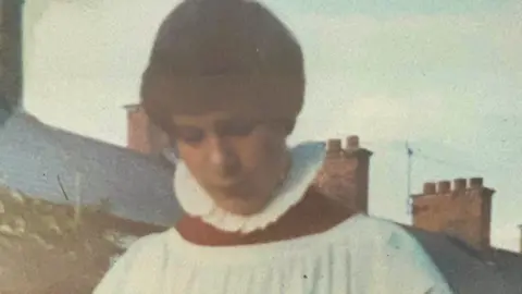 A young boy dressed in a white choir surplice. It is frilled around the neck and has a large red collar. The boy, who has dark hair, is looking down towards the ground. The background of the grainy image shows a blue sky and red bricked chimneys along a slate roofline. 