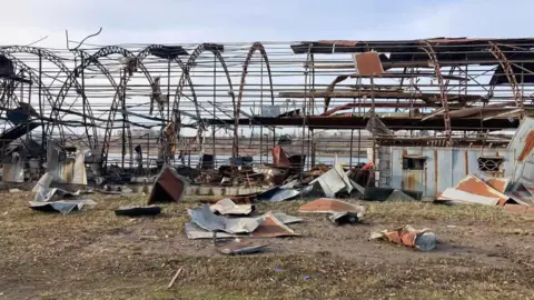 Odesa Dragons Image shows the Odesa Dragon boathouse after it was destroyed in an attack. The metal framing is still standing but the panelling has completely come off of it. Metal panels are laying on the floor around the structure. Behind the structure, the sky is blue. There is water behind the structure too.