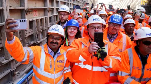 PA Media Men and women in orange high-vis jackets are standing with their phones out. They have white hard hats on that read "HS2" in blur writing. They are wearing protective goggles.