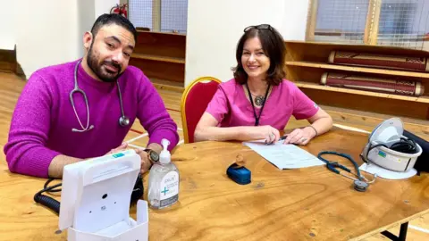Swansea Bay University Health Board Mootaz and Wynn are both sat at a wooden desk inside the mosque with medical equipment and paperwork in front of them. Mootaz is wearing a long-sleeved purple top and has a stethoscope around his neck. He has short dark hair and dark facial hair. Wynn is wearing a pink short sleeved top and has shoulder-length dark hair, and is wearing glasses on top of her head. Both are smiling at the camera.