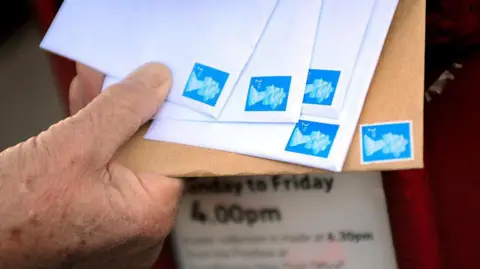 Close up of the hand of a man posting five letters with blue second-class stamps on in to a post box