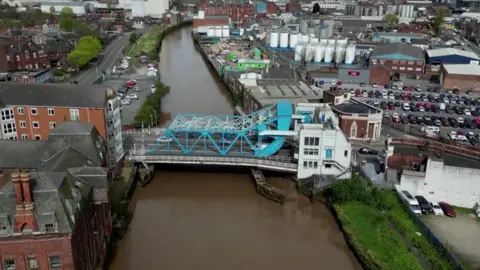 An aerial view of a blue and white bridge spans over a brown river. Industrial looking buildings, tower blocks and car parks are visible on either side of the bank.