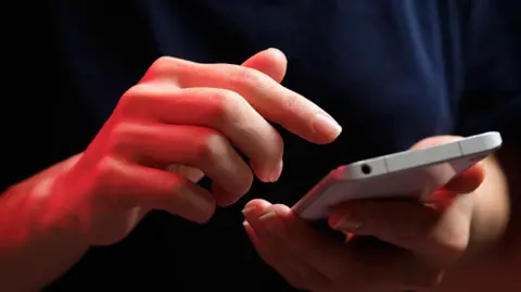 A woman's hands with a mobile phone in her left hand, with her right hand being used to hover over it before she presses it.