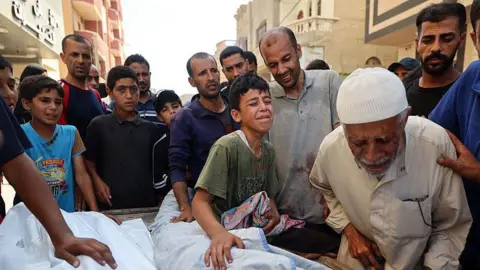 A group of men and boys stand around a child in a body bag, some crying and touching the shroud, during a funeral on the street in Gaza.