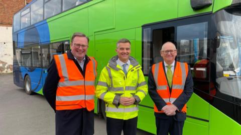 Gloucestershire County Council leader Cllr Mark Hawthorne, Transport Secretary Mark Harper and Cllr Philip Robinson at the Cheltenham depot. The three men are shown wearing high-vis clothing, in front of a bright green bus. 