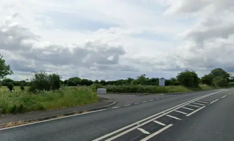 LDRS A field next to a road with a turning into the field blocked with concrete blocks and a 'for sale' sign next to the field