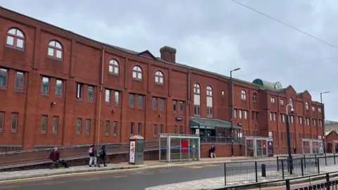 A wide red brick building with three bus stops in front of it