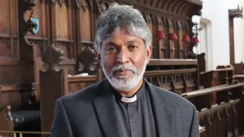 Priest Isaac Poobalan - a man with grey hair and a beard, a dog collar and a dark grey jacket, stands in a church, with several rows of choir stalls behind him.