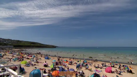 BBC A photo of Porthmeor beach. It shows hundreds of people on the beach, with some people swimming and paddling in the sea. There are towels, tents and umbrellas on the beach, with people sunbathing. The sky is blue.
