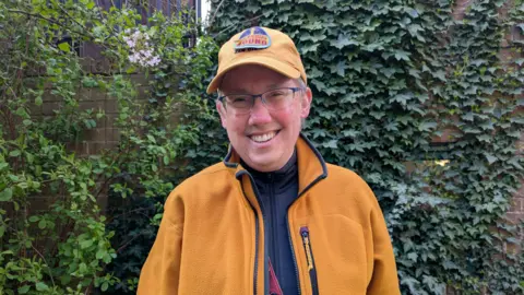 Julie Mariotti/BBC A participant in the guided Anne Lister tour standing in front of an ivy wall in the Holy Trinity Church's garden with a big smile on their face.