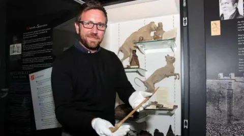 Chris Morris Daniel Clarke smiles at the camera holding a witches wand in front of an exhibition. He has short dark hair and he wears black glasses with a black jumper. He wears white gloves.