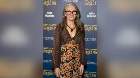 Craig Sugden Rachel Joyce stands in front of signage promoting the press night of The Unlikely Pilgrimage of Harold Fry. She has long grey hair, glasses and wears a dress with orange flowers and lace sleeves. 