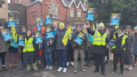 MICHELLE GREAVES A group of teachers with signs saying Pay Up stand in front of a red brick school building. 