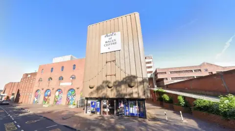 A large building with a ramp at the side of it. There is a sign which reads Prince of Wales Centre above a set of windows and glass doors. A section of the building set back from the entrance has a number of archways which each contain colourful murals.
