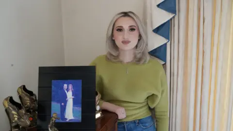 John Fairhall/BBC A woman, wearing a green top and blue jeans, with long blonde hair and wearing a necklace, stands in front of a pair of curtain. A picture frame of a man and a woman is in front of her positioned on a cabinet. She is leaning one arm on the cabinet. 