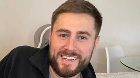 a man with short brown hair and a short brown beard, wearing a black fleece top and grey t-shirt, smiles for the camera