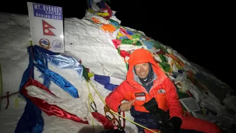 A climber wearing a bright orange coat and trousers smiles as he sits on top of Everest at night. He looks very cold and is connected to ropes as he sits against a snowy bank, which has a summit marker with the Nepal flag and "Mount Everest Spring 2026" on it.