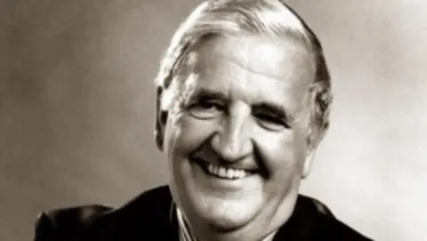 A black and white image of Sir Kenneth Bloomfield smiling at the camera as he wears a suit.