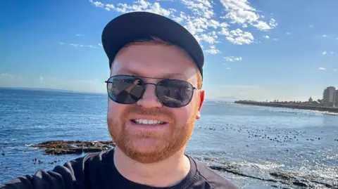 Ulrich Janse van Vuuren, with a cap and sunglasses, overlooking the ocean in South Africa