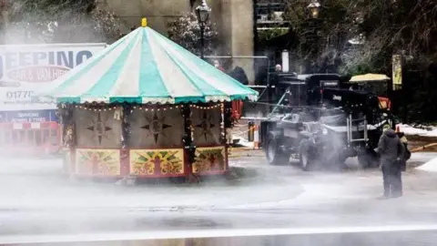 A fairground stall can be seen next to St Albans' Verulamium lake as fake snow is sprayed around. A person is standing by a vehicle, there are vans, a street lamp with snow on it and a colourful fairground stall. 