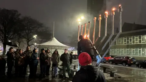 Rabbi Mendy Singer stands in front of the menorah to light the candles. He is on a step ladder as people look on.