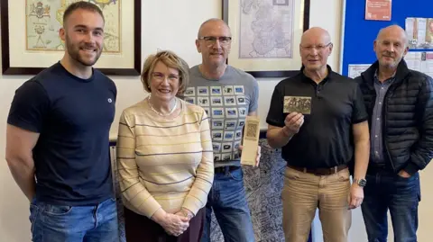 Philipp Cross A young man in his 20s wears a blue t-shirt and jeans, standing next to one elderly woman and three older men at a library. Two men in the middle are holding WW1 postcards