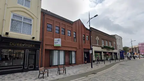 Empty red brick building on a high street. It has a To Let sign on the front, and is flanked by a pub and a shop