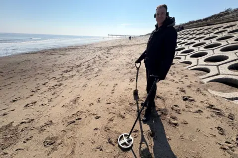 Kevin Shoesmith/BBC Chris Morris berdiri di pantai Skegness dengan detektor logam di depan dinding laut yang miring. 