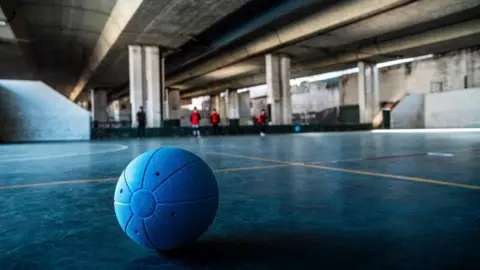 Getty Images A close-up of a ball for visually impaired people on a court. The ball is blue with holes in and players are on the other side of the room in the background.