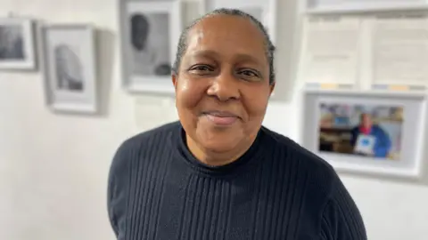 Pauline Swaby-Wallace has short black hair and is wearing a black top. she is smiling at the camera and has brown eyes. In the background is the photography exhibition.