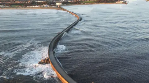 A pier with a road on it stretches out into the blue ocean. There are rocks dotting the border between the pier and the water. The town of South Shields can be seen in the background.