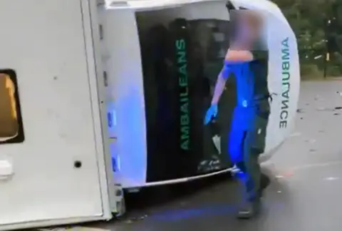 BBC A paramedic is seen next to an overturned ambulance 