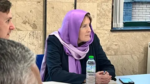 Emma Baugh/BBC Sarah, a women wearing a purple headscarf and a black jacket is sitting on a table addressing the meeting.