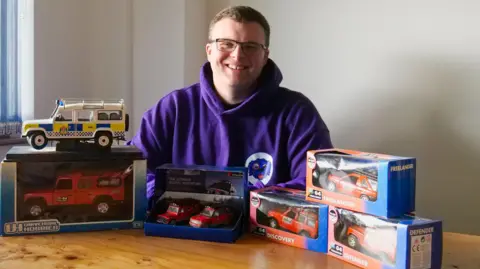 Elliot Riley-Walsh Elliot wearing a purple hoodie and glasses sits at a wooden table displaying a collection of scale-model Land Rover vehicles. The collection includes a white emergency-service Land Rover on a display case, several red Land Rover models in branded packaging, and boxed models labelled Discovery, Freelander, and Defender. The scene is lit by natural light from a window on the left, with a plain light-coloured wall in the background.