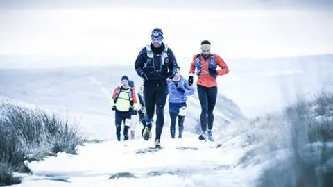 Montane Cheviot Goat A group of runners appear on the brow of a hill. There is snow on the ground and they are all wrapped up in warm running gear.  