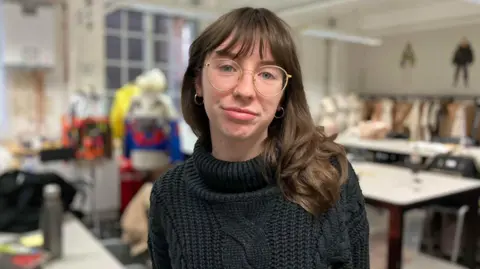 Alex Dunlop/BBC Ruby Ormston is wearing a dark knitted sweater and has shoulder‑length wavy hair and gold-rimmed glasses. In the background, the studio is filled with large tables, sewing equipment, and mannequins draped in colorful material. 