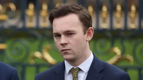 PA Media Eoin Tennyson - a man with short, brown hair is standing in front of an black and gold iron gate. He is wearing a dark suit jacket, a white collared shirt and a pattern yellow tie.