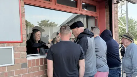 Three people are behind the window serving people outside with tickets. There are five men facing the window.