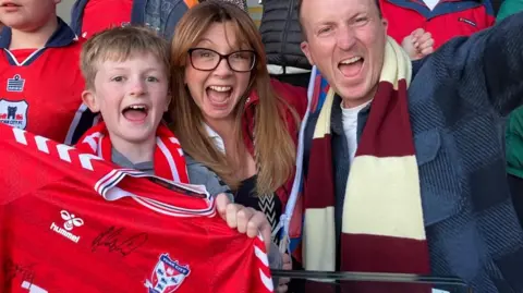A woman, man and young boy smile at the camera. The boy is holding a York City top with an autograph on it.