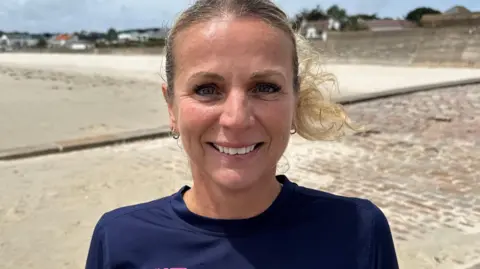 A blonde-haired woman stood on a path beside a sandy beach. 